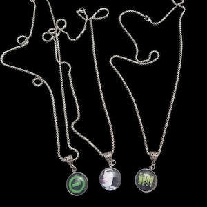 May include: Three silver-toned necklaces with round pendants. Each pendant features a different image: a green checkmark, a portrait, and a band photo. The necklaces are displayed against a black background.