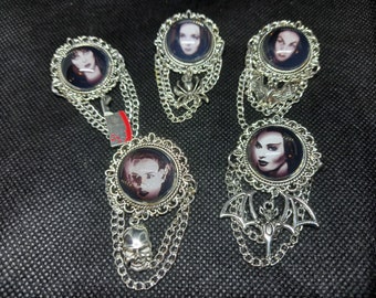 Op de afbeelding: Vijf zilveren broches met zwart-wit portretten van vrouwen. Elke broche heeft een ander ontwerp, waaronder kettingen, schedels en vleermuisvleugels.