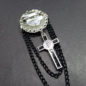 Op de afbeelding: Zilverkleurige broche met een cirkelvormig ontwerp, versierd met heldere strass-steentjes. Een zwarte ketting hangt van de broche, met een zilveren kruis hanger met zwarte accenten. Het kruis heeft een religieus embleem.