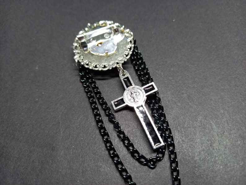 Op de afbeelding: Zilverkleurige broche met een cirkelvormig ontwerp, versierd met heldere strass-steentjes. Een zwarte ketting hangt van de broche, met een zilveren kruis hanger met zwarte accenten. Het kruis heeft een religieus embleem.