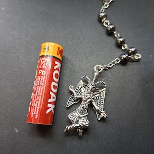 K&ouml;nnte beinhalten: Ein silberfarbener Metallanh&auml;nger in Form einer geh&ouml;rnten Figur mit Fl&uuml;geln, befestigt an einer Kette mit dunklen Metallperlen. Eine rot-gelbe Kodak AAA-Batterie liegt neben dem Anh&auml;nger, um die Gr&ouml;&szlig;e des Schmuckst&uuml;cks im Vergleich zu verdeutlichen.