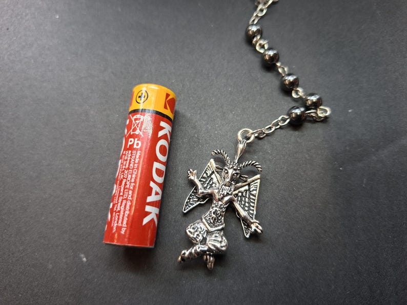 K&ouml;nnte beinhalten: Ein silberfarbener Metallanh&auml;nger in Form einer geh&ouml;rnten Figur mit Fl&uuml;geln, befestigt an einer Kette mit dunklen Metallperlen. Eine rot-gelbe Kodak AAA-Batterie liegt neben dem Anh&auml;nger, um die Gr&ouml;&szlig;e des Schmuckst&uuml;cks im Vergleich zu verdeutlichen.