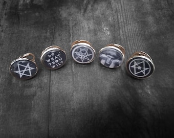 May include: Five circular rings with rose gold-toned metal bands and glass cabochon tops. The designs include white geometric star symbols on black backgrounds, the text DO WHAT THOU WILT, and a black and white portrait of a person with hands.