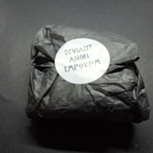 May include: A package wrapped in black tissue paper, secured with a white circular sticker. The sticker features the text "DEVIANT ANGEL EMPORIUM" in a stylized font. The background is a solid black surface.