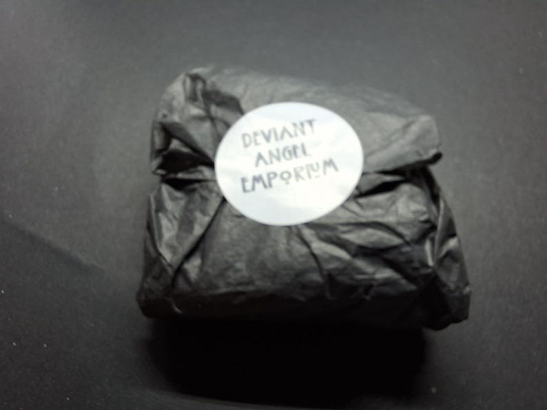 May include: A package wrapped in black tissue paper, secured with a white circular sticker. The sticker features the text "DEVIANT ANGEL EMPORIUM" in a stylized font. The background is a solid black surface.