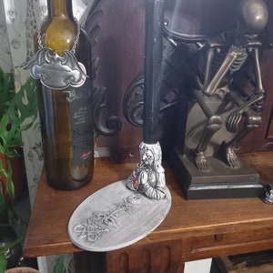 May include: A black and white figurine of a woman with a red nose, holding a black candle. The figurine is on a gray oval base with the word "remarkable" written on it. The figurine is on a wooden table with a bottle of wine and a skeleton figurine in the background.