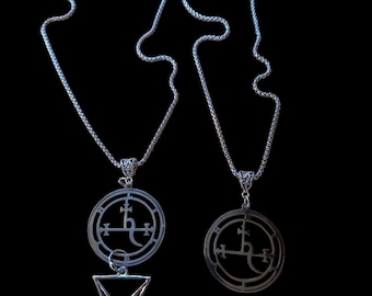May include: Two silver-toned necklaces featuring circular pendants with intricate etched sigil designs. One pendant is paired with a dangling triangular symbol featuring a cross-like base. Both necklaces have textured silver chains.