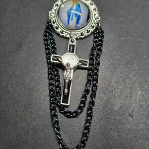 Op de afbeelding: Zilverkleurige broche met een religieuze afbeelding in een decoratief frame. Een zwarte kruis hanger met een figuur van Jezus hangt eronder, met zwarte ketting accenten.