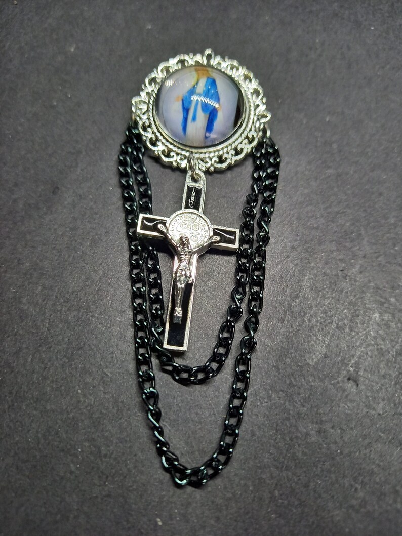 Op de afbeelding: Zilverkleurige broche met een religieuze afbeelding in een decoratief frame. Een zwarte kruis hanger met een figuur van Jezus hangt eronder, met zwarte ketting accenten.