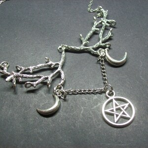 May include: Silver-toned necklace featuring a branch-like design with two crescent moons and a pentagram pendant. The necklace has a delicate chain and is set against a dark background. The jewelry is likely made of metal.