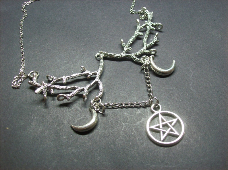 May include: Silver-toned necklace featuring a branch-like design with two crescent moons and a pentagram pendant. The necklace has a delicate chain and is set against a dark background. The jewelry is likely made of metal.