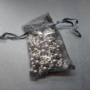 K&ouml;nnte beinhalten: Ein schwarzer, transparenter Organza-Beutel mit Kordelzug, der eine silberfarbene Kettenhalskette mit kleinen Metallperlen und einem dreieckigen Anh&auml;nger enth&auml;lt. Der Beutel besteht aus feinem, halbtransparentem Netzstoff mit passenden schwarzen B&auml;ndern.