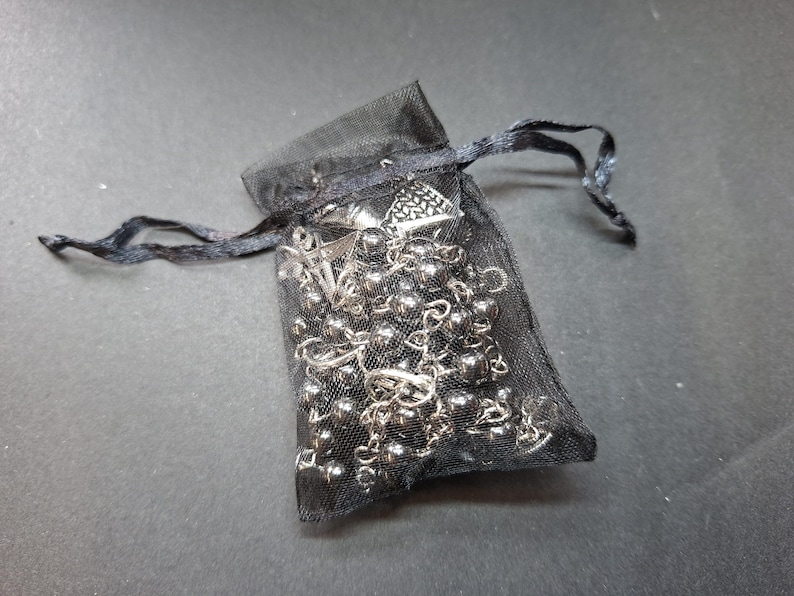 K&ouml;nnte beinhalten: Ein schwarzer, transparenter Organza-Beutel mit Kordelzug, der eine silberfarbene Kettenhalskette mit kleinen Metallperlen und einem dreieckigen Anh&auml;nger enth&auml;lt. Der Beutel besteht aus feinem, halbtransparentem Netzstoff mit passenden schwarzen B&auml;ndern.