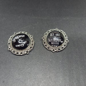 May include: Two silver-toned brooches with black glass cabochons. The left brooch features the text "Strange & Unusual" in white. The right brooch features the text "We are the Weirdos Mister" in white.