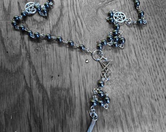 Könnte beinhalten: Eine Perlenkette mit dunklen, metallisch glänzenden, kugelförmigen Perlen, die durch silberfarbene Metallketten verbunden sind. Die Kette enthält mehrere silberfarbene Pentagramm-Anhänger, ein Siegel-Symbol und ein umgekehrtes Kreuz.