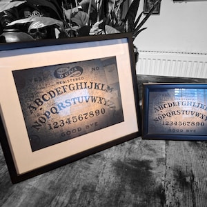 Può includere: Due stampe di tavola Ouija in cornici nere. La stampa più grande a sinistra presenta un passe-partout color crema, mentre quella più piccola a destra ne è priva. Entrambe mostrano l'alfabeto, i numeri da zero a nove e la scritta GOOD BYE.