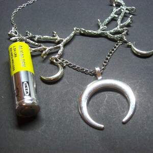 May include: Silver-toned necklace featuring a crescent moon pendant and smaller moon charms. The chain is adorned with a branch-like design. An IKEA AA alkaline battery is placed next to the necklace.