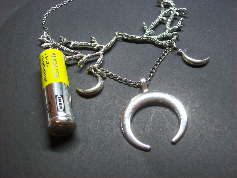 May include: Silver-toned necklace featuring a crescent moon pendant and smaller moon charms. The chain is adorned with a branch-like design. An IKEA AA alkaline battery is placed next to the necklace.