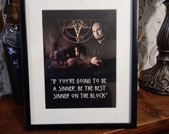 May include: A black framed print with a quote that reads "IF YOU'RE GOING TO BE A SINNER, BE THE BEST SINNER ON THE BLOCK". The print features a man in a priest's collar standing in front of a pentagram.