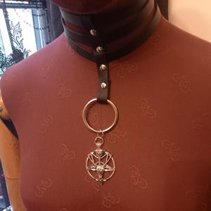 May include: A black leather choker necklace with a silver pendant. The choker has a layered design with silver rivets. The pendant is a silver pentagram with a goat head in the center, suspended from a large silver ring.