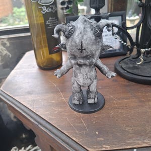 May include: A small, grey, stone-textured figurine of a horned creature with cloven hooves and a black inverted cross symbol on its forehead. The figure stands on a circular black base with its arms slightly extended to the sides.