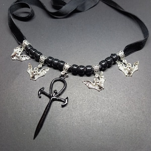 May include: Black velvet choker necklace with black beads and silver bat charms. The necklace also features a black ankh pendant.