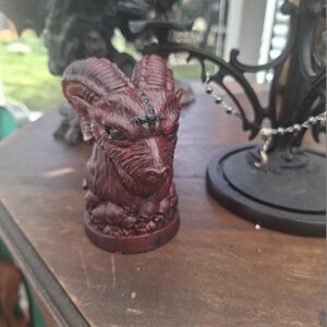 May include: A dark red, sculpted bust of a horned goat head featuring a black inverted cross on the forehead. The base of the sculpture is decorated with a cluster of small, detailed skulls, and the entire piece has a textured, weathered finish.