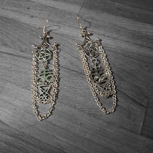 May include: A pair of gold-colored dangle earrings. Each earring features a decorative top with a pentagram, a symbol, and chain details. The earrings are displayed on a gray wooden surface.