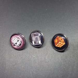 Könnte beinhalten: Drei runde Metall-Pins mit verschiedenen Designs. Der erste Pin zeigt ein pinkfarbenes und weißes Bild eines Charakters aus dem Film Hellraiser. Der zweite Pin hat ein schwarz-weißes Design mit dem Text "STRAIGHT OUTTA HELL". Der dritte Pin hat einen schwarzen Hintergrund mit einem goldenen Bild eines Würfels.