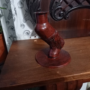 Puede incluir: Un portavelas de madera con forma de pierna estilizada con una superficie texturizada. El portavelas es de color marrón rojizo oscuro y tiene una base redonda.