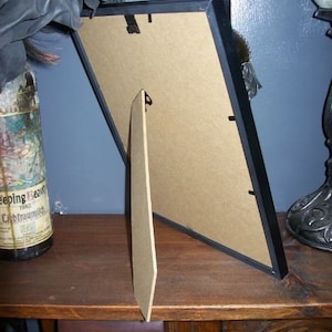 May include: A black picture frame with a cardboard backing and a wooden easel stand. The frame is angled, revealing the back and stand. A bottle with a label is partially visible on the left.