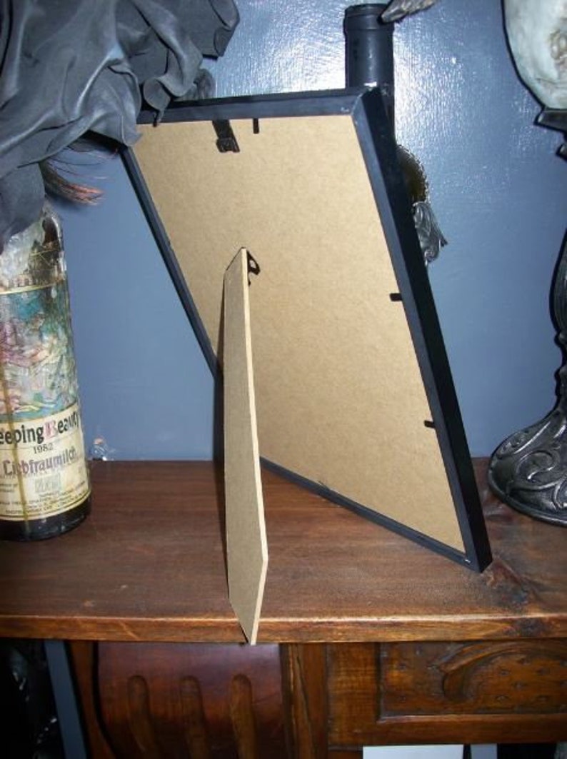 May include: A black picture frame with a cardboard backing and a wooden easel stand. The frame is angled, revealing the back and stand. A bottle with a label is partially visible on the left.