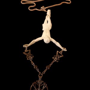 Peut inclure: Un collier pendentif avec une figurine crème suspendue à une chaîne dorée. La figurine est reliée à une chaîne avec un pendentif pentagramme et deux triangles inversés. Le fond est noir.