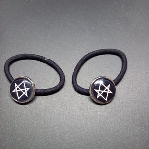 Könnte beinhalten: Zwei schwarze Haargummis mit silberfarbenen runden Anhängern, die ein weißes Pentagramm zeigen.