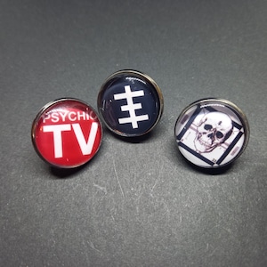 Puede incluir: Tres pines redondos de metal con diferentes diseños. El primer pin tiene un fondo rojo con texto blanco que dice "PSYCHIC TV". El segundo pin tiene un fondo negro con un símbolo blanco. El tercer pin tiene un fondo blanco con un dibujo en blanco y negro de una calavera.