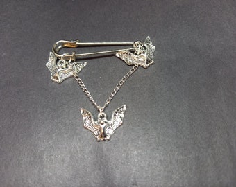 Op de afbeelding: Een zilverkleurige veiligheidsspeld-broche met drie kleine vleermuisvormige bedels. Twee vleermuizen zijn direct aan de speld bevestigd, terwijl de derde aan een fijn zilveren kettinkje tussen de twee bovenste bedels hangt.