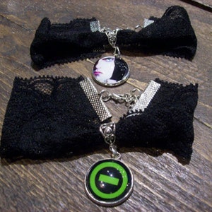 Könnte beinhalten: Zwei schwarze Spitzen-Choker-Halsketten mit silbernen Metallakzenten. Ein Anhänger zeigt ein Frauen Gesicht, der andere einen grünen Kreis mit einer schwarzen Linie.