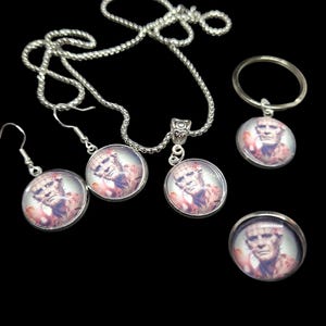 May include: A jewelry set featuring a pendant necklace, earrings, a keychain, and a ring. Each piece has a silver-toned setting and a circular image of a person. The background is black.