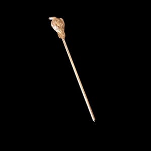 以下が含まれることがあります： 彫刻された鳥の飾りが付いたゴールドトーンの金属製ヘアピン。鳥は止まっている姿勢で、翼と羽の質感が細かく表現されています。ピンは長く細い直線の軸で、先端に向かって細くなっています。