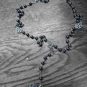 K&ouml;nnte beinhalten: Eine Perlenkette mit dunklen, metallisch gl&auml;nzenden, kugelf&ouml;rmigen Perlen, die durch silberfarbene Metallketten verbunden sind. Die Kette enth&auml;lt mehrere kreisf&ouml;rmige silberfarbene Pentagramm-Anh&auml;nger und einen zentralen silberfarbenen Anh&auml;nger mit einer gefl&uuml;gelten Figur.