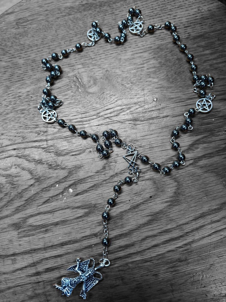 K&ouml;nnte beinhalten: Eine Perlenkette mit dunklen, metallisch gl&auml;nzenden, kugelf&ouml;rmigen Perlen, die durch silberfarbene Metallketten verbunden sind. Die Kette enth&auml;lt mehrere kreisf&ouml;rmige silberfarbene Pentagramm-Anh&auml;nger und einen zentralen silberfarbenen Anh&auml;nger mit einer gefl&uuml;gelten Figur.
