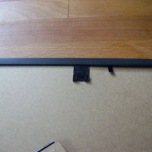 May include: A black picture frame with a cardboard backing. The frame is rectangular with a thin, black border. The back has a metal hanger and a small metal clip to hold the backing in place. The frame is on a wooden surface.
