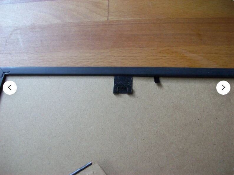 May include: A black picture frame with a cardboard backing. The frame is rectangular with a thin, black border. The back has a metal hanger and a small metal clip to hold the backing in place. The frame is on a wooden surface.