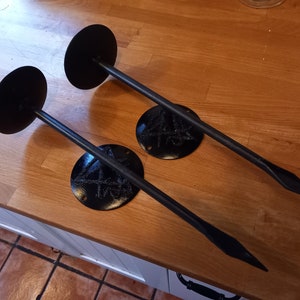 May include: Two black metal wall mounted candle holders with a pointed tip on each arm. Each holder has a round base with a decorative design.