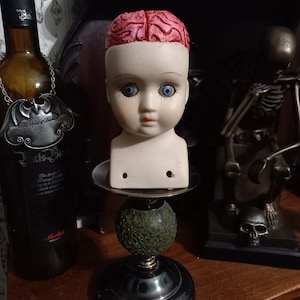 Puede incluir: Una cabeza de muñeca con ojos azules y una parte superior en forma de cerebro rosa se encuentra sobre un candelabro decorativo. El soporte tiene una placa plateada, una esfera de mosaico verde y una base negra. Una botella de vino y una figura de esqueleto están en el fondo.