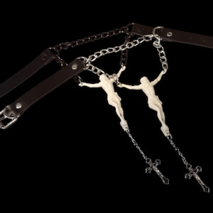May include: A dark brown leather and metal bondage accessory. Two white crucifix figures hang from silver chains. The accessory has two leather straps with silver buckles and a chain that connects the two straps.