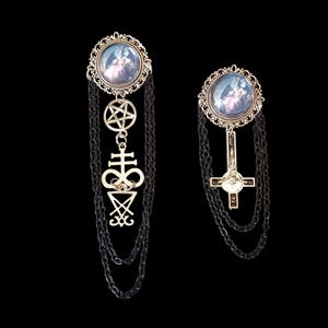 Peut inclure: Deux broches gothiques avec des cadres argentés ornés et des chaînes noires. L'une présente un pentagramme et un pendentif sigillaire, l'autre une croix. Chaque broche a une image circulaire avec une scène mystique.