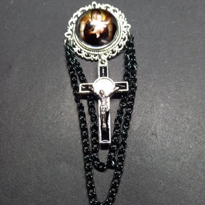 Op de afbeelding: Zilverkleurige broche met een religieuze afbeelding in een decoratief frame. Een zilveren kruis met een figuur van Jezus hangt eronder, met zwarte kettingaccenten. De broche is geplaatst tegen een donkere achtergrond.