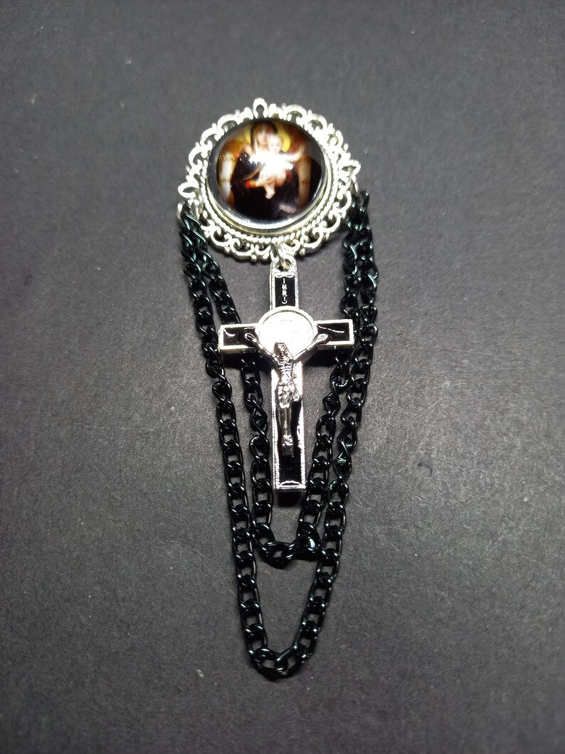 Op de afbeelding: Zilverkleurige broche met een religieuze afbeelding in een decoratief frame. Een zilveren kruis met een figuur van Jezus hangt eronder, met zwarte kettingaccenten. De broche is geplaatst tegen een donkere achtergrond.