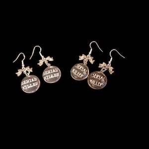 May include: A pair of silver earrings with a small bow charm and a round glass charm that says "SERIAL KILLER" in white lettering.